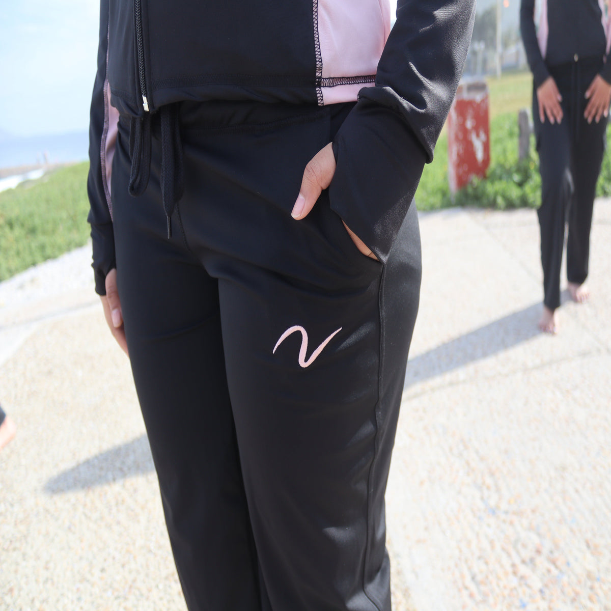 A person wearing black athletic jogger pants with a pink script logo on the thigh, standing outdoors on a paved path. The pants have side pockets and a drawstring waist.