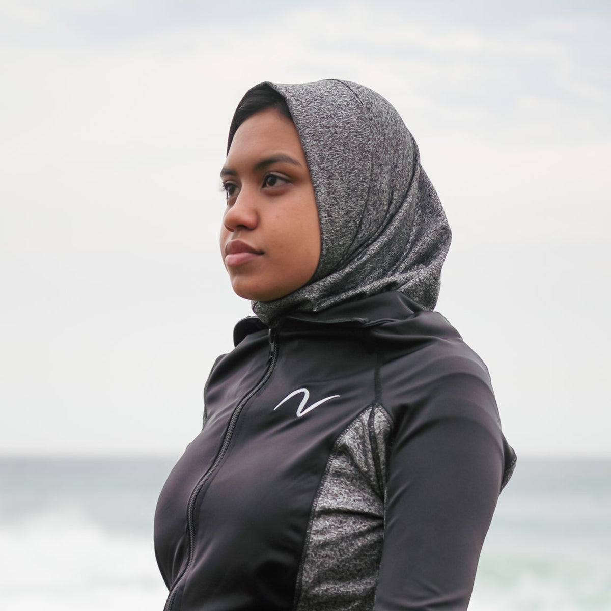 A woman wearing a black and gray athletic zip-up hoodie with a matching hijab and black athletic pants, posing outdoors with a beach and ocean background.