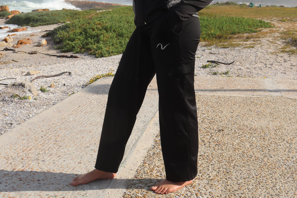 A woman stands barefoot on a gravel path outdoors near the ocean, wearing a black long-sleeve top and matching black pants with a small white stylized logo on the chest and upper thigh.