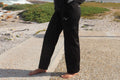 A woman stands barefoot on a gravel path outdoors near the ocean, wearing a black long-sleeve top and matching black pants with a small white stylized logo on the chest and upper thigh.