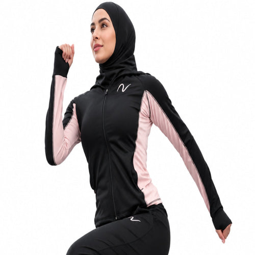 A woman wearing a black and pink athletic hijab set with black pants and white sneakers is captured mid-jump on an outdoor path with a rocky cliff in the background.
