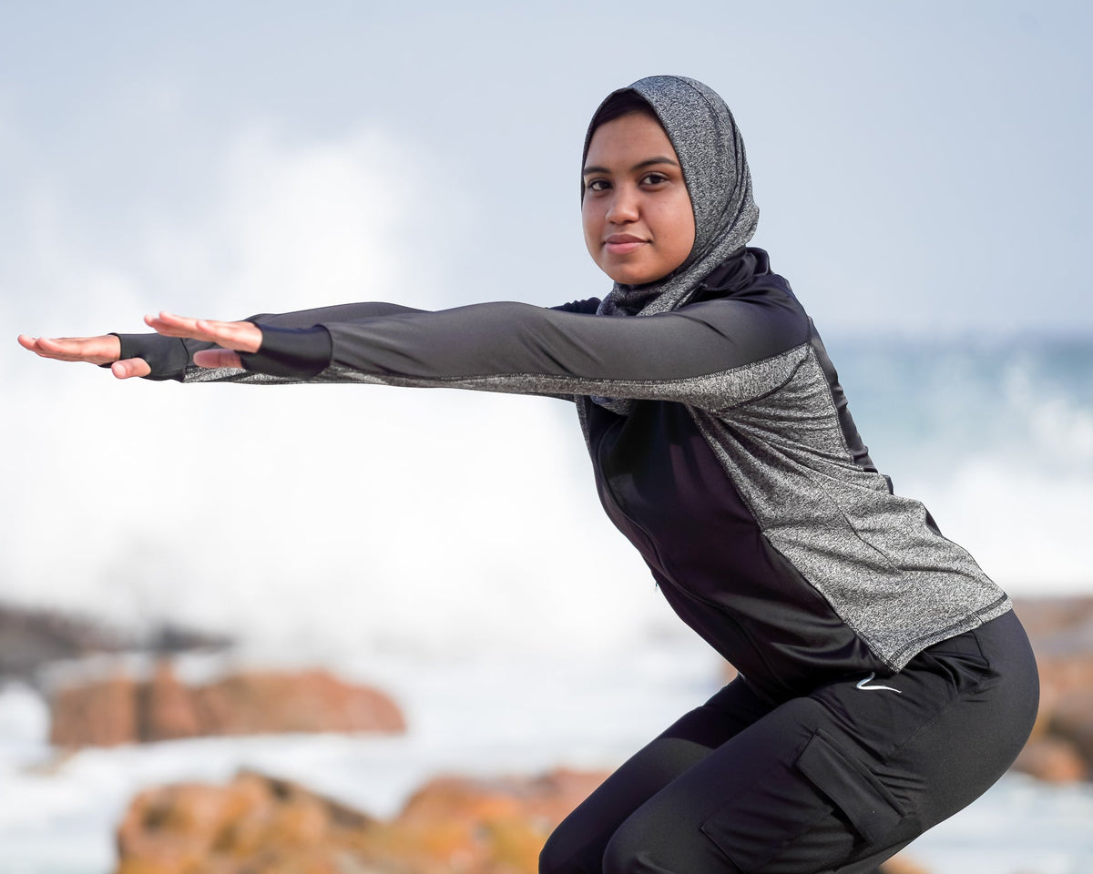 A woman in a gray and black athletic hijab and matching long-sleeve top poses in a crouched yoga or stretching pose on a rocky beach with ocean waves in the background.