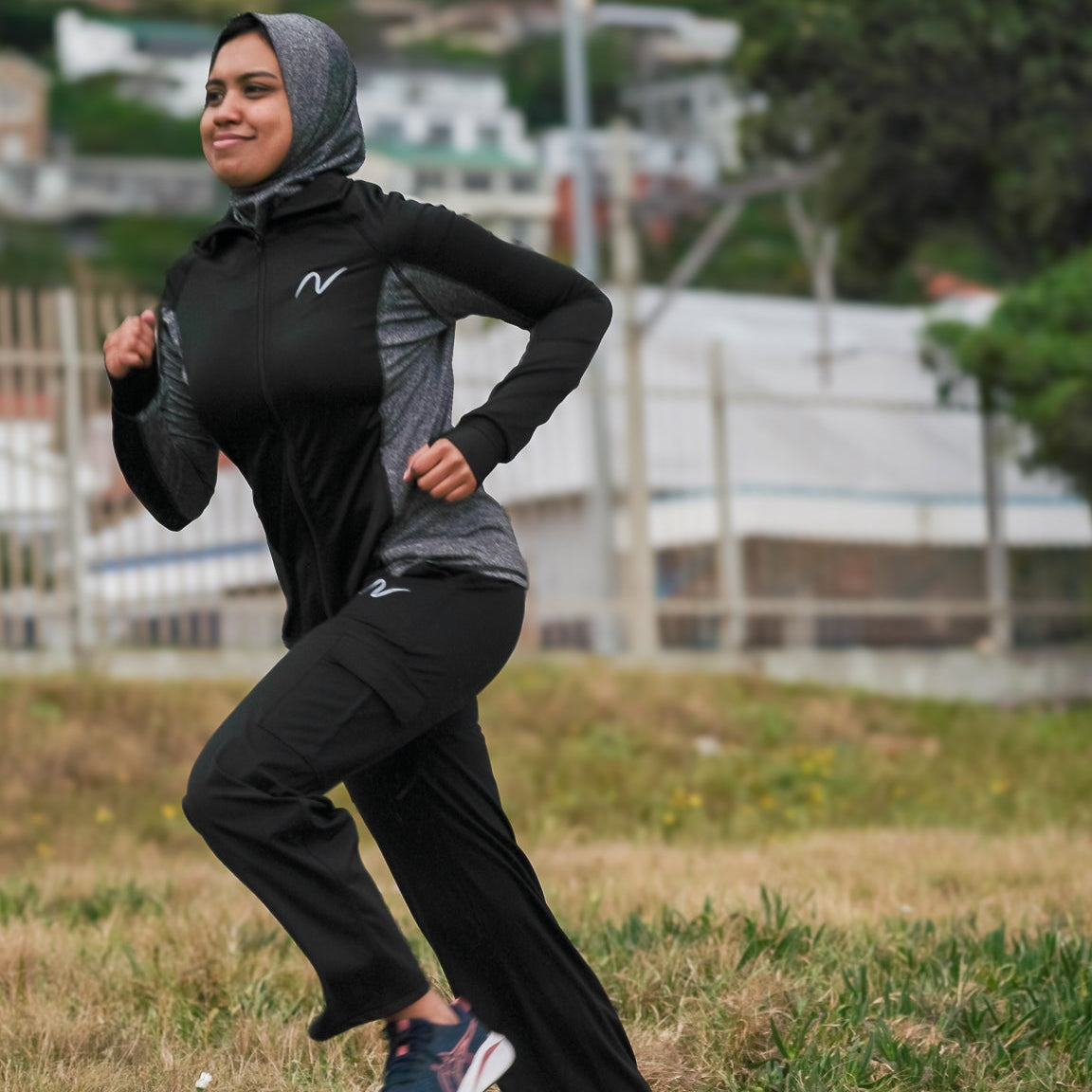 A woman wearing a black and gray long-sleeve athletic hoodie with a matching hijab and black running pants is jogging on an outdoor paved path. The background shows grass, trees, and buildings in a park setting.