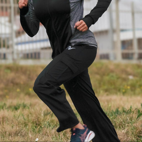 A woman wearing a black and gray long-sleeve athletic hoodie with a matching hijab and black running pants is jogging on an outdoor paved path. The background shows grass, trees, and buildings in a park setting.