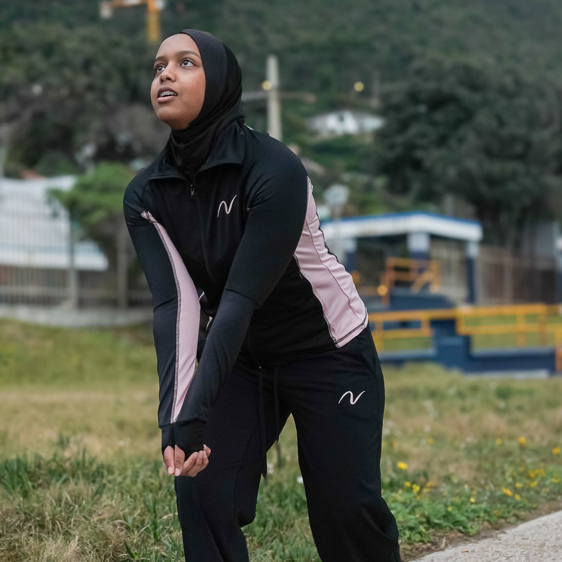 A woman wearing a black hijab and a matching black athletic tracksuit with pink side panels and white 'N' logos, posing outdoors on a paved path with a mountain and crane in the background.