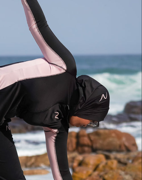 A woman in a black and light pink long-sleeve activewear set with matching black pants is stretching on a rocky beach with the ocean and sky in the background.