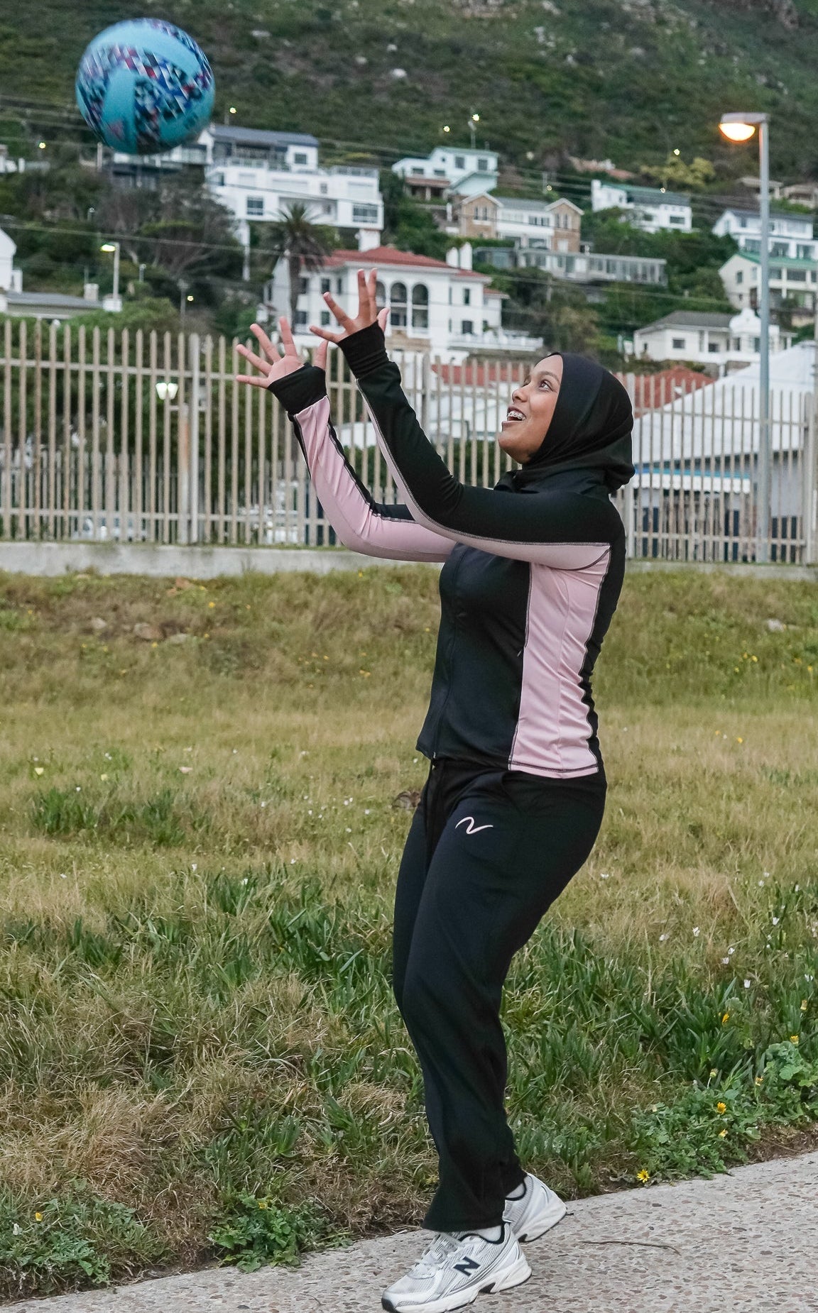 A woman in a black hijab and a black and pink athletic tracksuit jumps to catch a blue patterned ball on a concrete path, with a grassy field, white fence, and houses on a hill in the background.