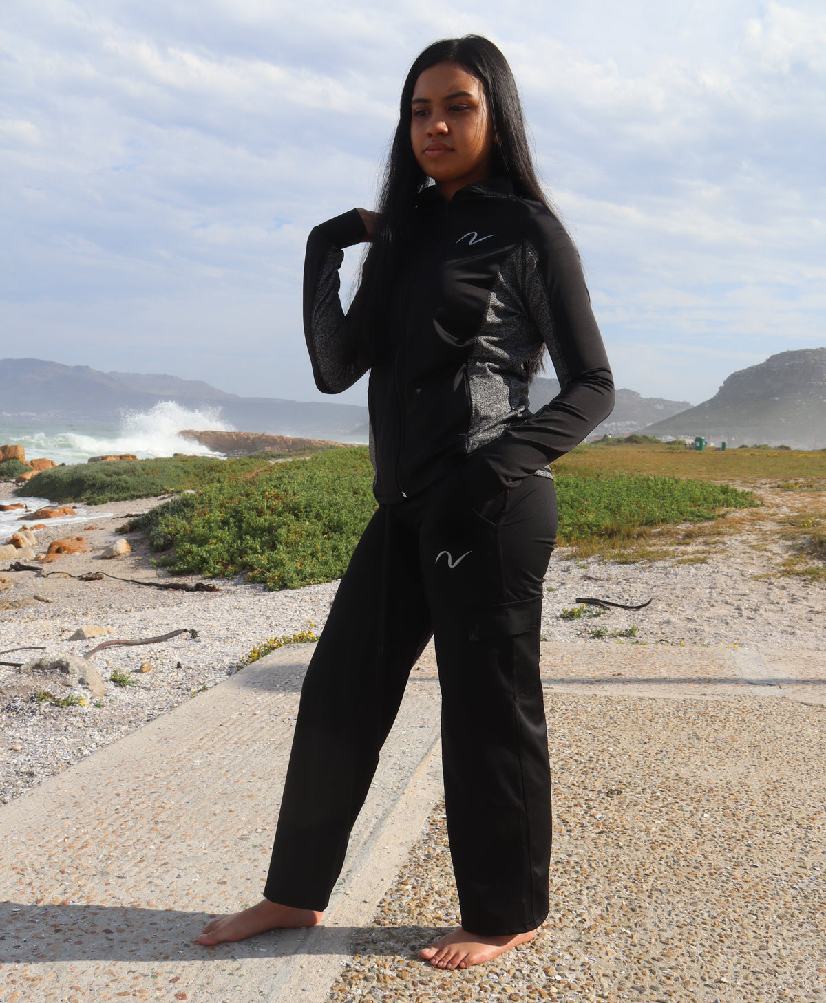 A woman stands barefoot on a gravel path outdoors near the ocean, wearing a black long-sleeve top and matching black pants with a small white stylized logo on the chest and upper thigh.